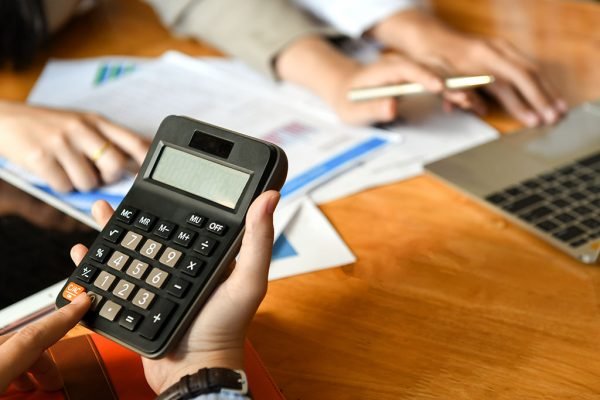 Imagem ilustrativa mostra duas pessoas utilizando calculadora, noteook e lidando com documentos em papel.
